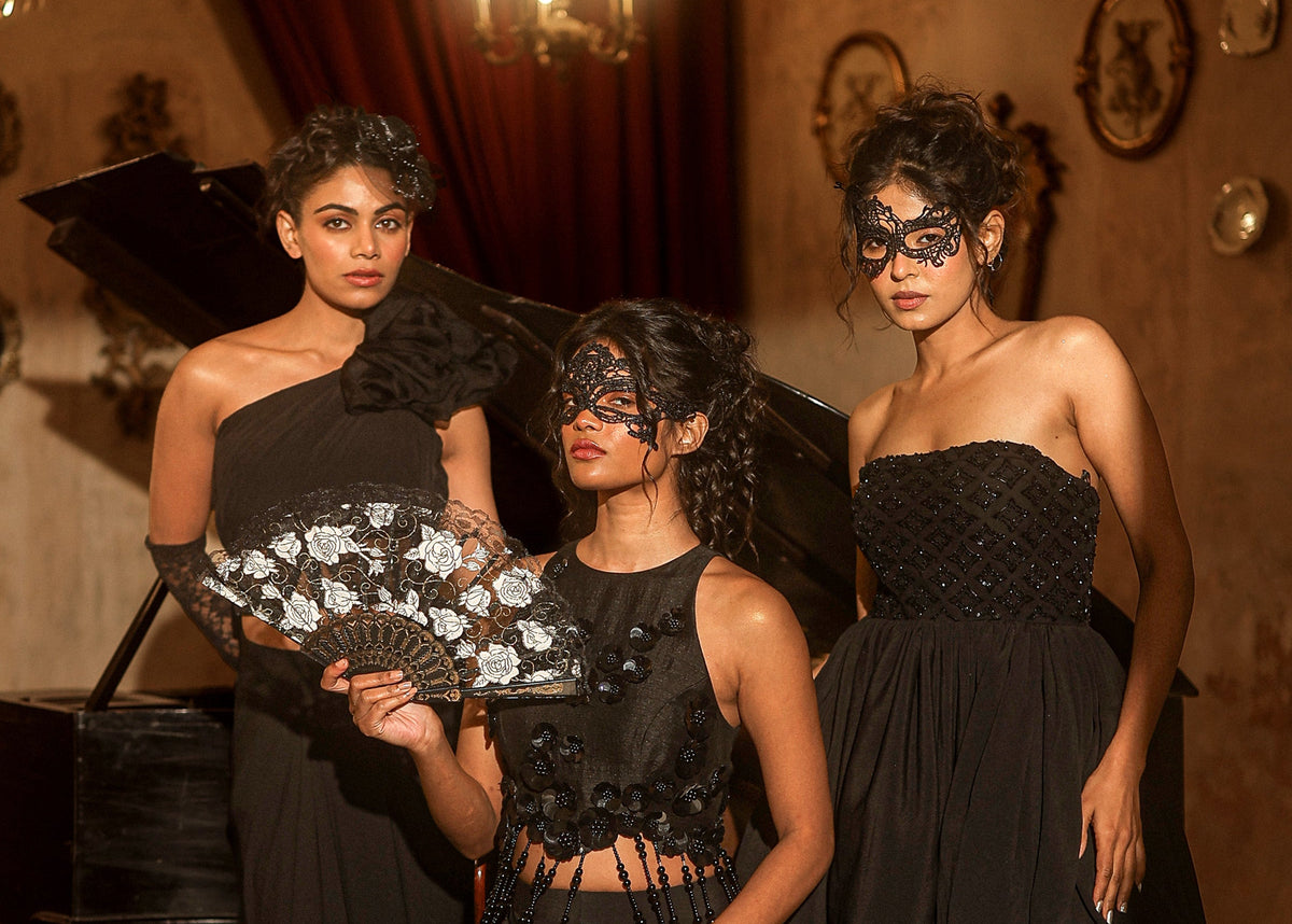 Mystery campaign: three models in black corset dresses, one with a lace fan and masquerade mask